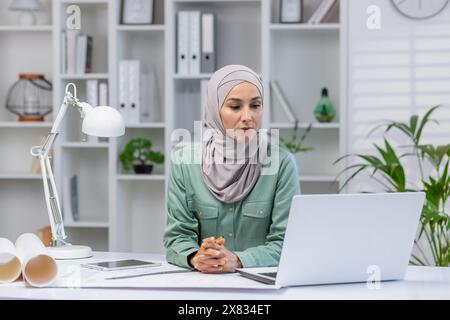 Woman in hijab designer working in home office, architect using laptop at work, studying technical assignment online, serious and concentrated reading. Stock Photo