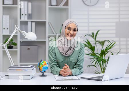 Portrait of happy teacher, woman in hijab sitting at desk smiling and looking at camera, muslim woman working remotely, teaching students and pupils using laptop in home office. Stock Photo
