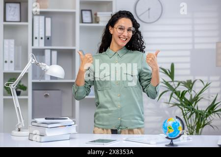 Portrait of a joyful female teacher in her own office, Latin American woman joyfully showing a thumbs up to the camera, teaching remotely, using a laptop. Stock Photo
