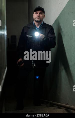 Mature male security guard in black uniform and cap walking upstairs in dark basement and lighting his way with flashlight Stock Photo