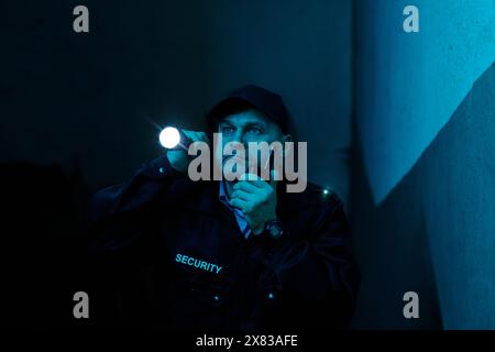 Serious security guard with walkie-talkie in hand notifying situation to his colleagues while lighting his way in dark corridor with flashlight Stock Photo