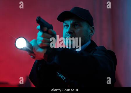 Mature man in cap and security uniform holding flashlight and gun while standing in dark corridor and looking straight during observation Stock Photo