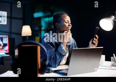 Excited black woman working remotely, using mobile device for video calls. Cheerful female blogger uses a laptop and cellphone to browse the internet, check emails, and stay connected. Stock Photo