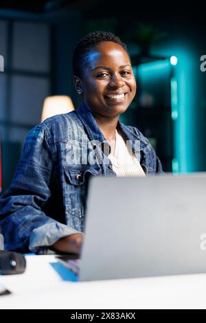 Portrait of African American blogger works on a laptop in a modern apartment. Smiling black woman is a dedicated freelancer, typing and reviewing information for her project. Stock Photo