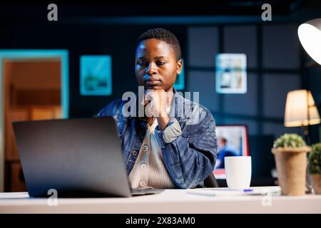 Female programmer sitting at home office desk and writing code on minicomputer, creating new software. African american woman working remotely, testing and looking for errors in program on laptop. Stock Photo