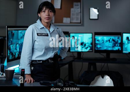 Young serious Hispanic female security guard in uniform looking at you while standing by workplace in surveillance room with cctv system Stock Photo