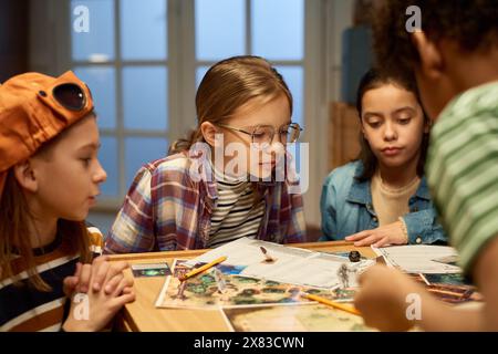 Group of friends looking at dice thrown by one of them while sitting by table and playing new leisure game with characters on board Stock Photo