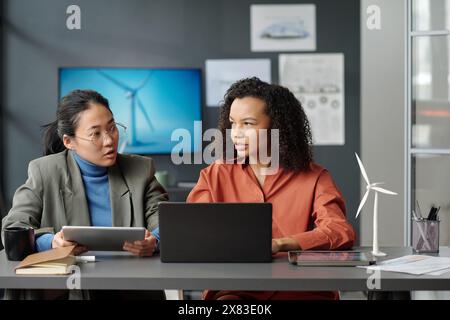 Two young successful intercultural female employees looking at one another while discussing online data and preparing new project Stock Photo