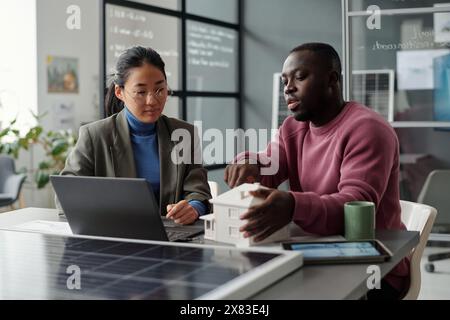 Two young intercultural architects looking at house model while discussing points of their new business project by workplace in office Stock Photo