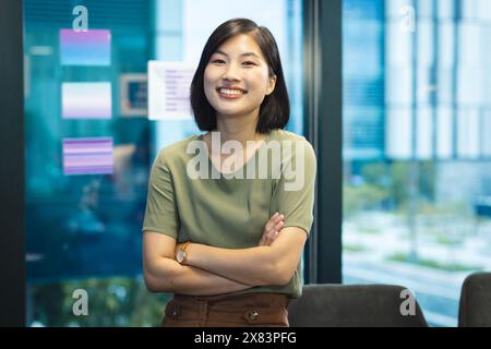 At modern office for business, young Asian woman standing with arms crossed, smiling confidently Stock Photo