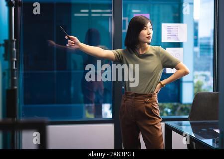 At modern office for business, young Asian woman standing, holding and pointing smartphone Stock Photo