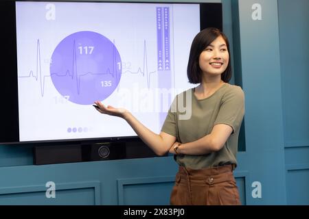 An Asian young woman in a modern office for business smiles and points at data on a screen Stock Photo