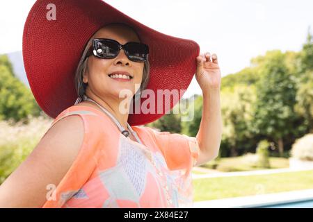 Asian senior female wearing large red hat and sunglasses, smiling outdoors Stock Photo