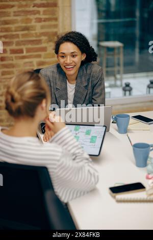 Two smiling female freelancers talking during work on laptop while sitting in cozy coworking Stock Photo