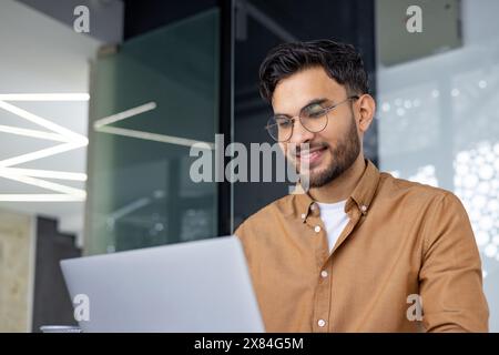 Smiling young man in a modern office using a laptop. Workspace environment with contemporary design. Stock Photo