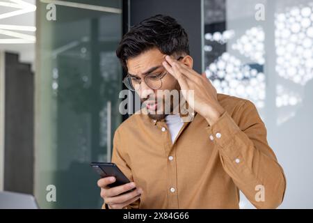 Young man in glasses looking shocked while reading something on his smartphone in a contemporary office environment Stock Photo