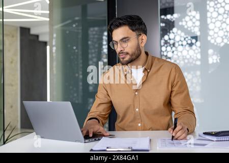 Focused businessman working on a laptop in a contemporary office, reviewing documents and making notes. Stock Photo