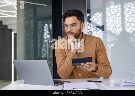 Focused professional working at a modern office desk, analyzing data on a laptop and holding a tablet. Business and technology concepts. Stock Photo