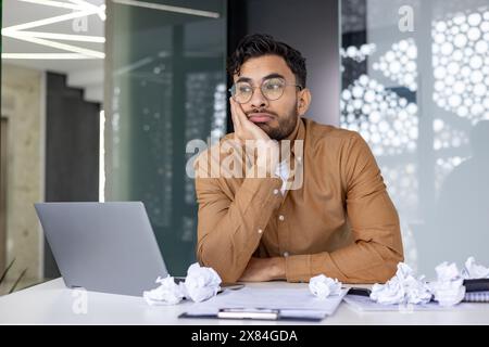 Frustrated young man in an office, looking away. He is sitting at a desk with a laptop surrounded by several crumpled papers. Stock Photo