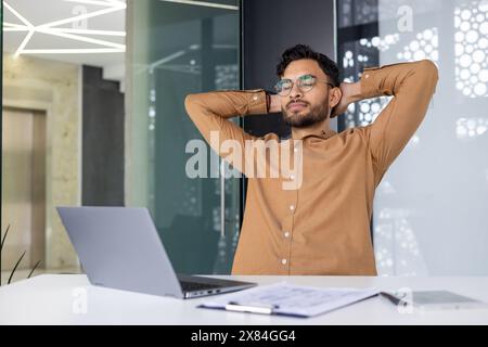 Confident businessman taking a break in modern office, feeling relaxed and accomplished. Man in glasses enjoying a moment of relaxation with hands behind head at his workplace. Stock Photo