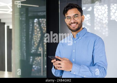 Portrait of a confident young man in a blue shirt smiling and holding a smartphone in a modern office setting. Stock Photo