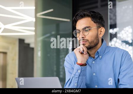 Young man in glasses and blue shirt concentrating while working on a laptop in a contemporary office space. Stock Photo