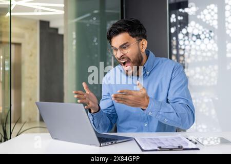 Man expressing frustration and anger at laptop in an office setting. Stress and business difficulties concept. Stock Photo