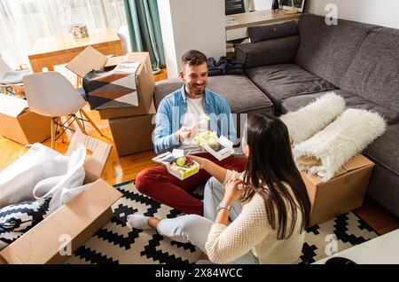 Smiling young couple move into a new apartment, sitting in the living room eating and taking a break from unpacking. Stock Photo