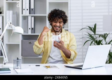 Excited young man celebrating success while looking at smartphone in a modern office setting. Achieving goals and experiencing happiness at work. Stock Photo