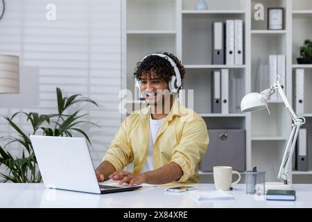 Man wearing headphones and working on a laptop in a comfortable, well-organized home office setting, emphasizing remote work and technology. Stock Photo