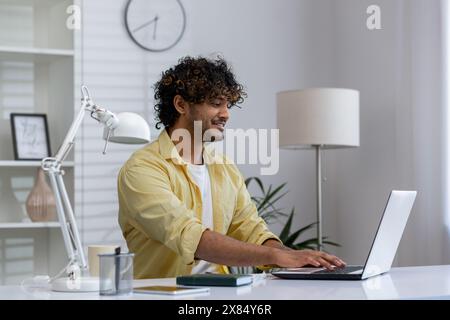 A cheerful young man in a yellow shirt working on his laptop at his home office desk. Modern, cozy workspace with natural light and minimalistic decor. Stock Photo