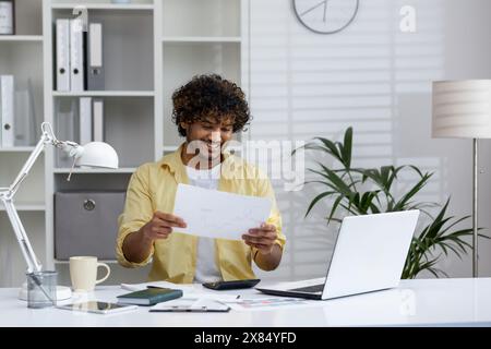 Smiling young man working from home at a desk with a laptop and documents. He appears happy and satisfied with his work. Stock Photo