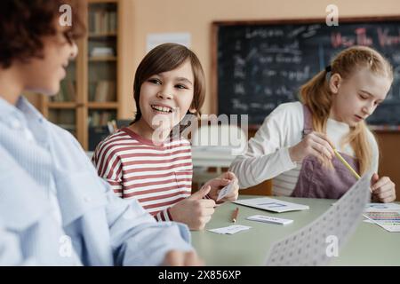 Cheerful Caucasian boy sitting at table in classroom chatting with his classmate during lesson Stock Photo