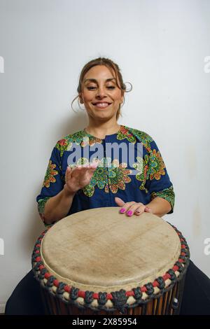 Portrait of an smiling happy Latin Woman playing the Yembe drum sitting at home. She is wearing an African shirt and has her hands on the drum leather Stock Photo
