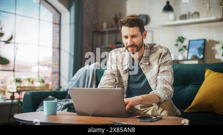 Portrait of Smiling Middle Aged Man Working from Home on a Laptop Computer in Sunny Cozy Apartment. Successful Male Entrepreneur Does Remote for e-Business Project, Online Shopping Stock Photo