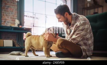 Happy Man Cuddles His Adorable Little Pug, Gives it Snacks at Home. Handsome Man Plays with His Dog, Best Friend, Scratches Super Happy Doggy, Have Fun in the Living Room Stock Photo