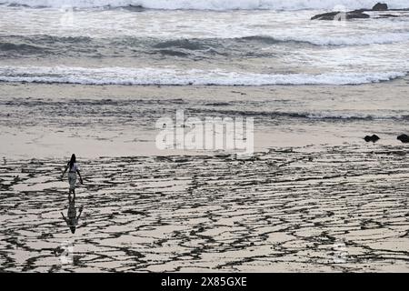 A woman walks along a beach with unique sand patterns created by the receding tide. The gentle waves in the background add to the serene and contempla Stock Photo