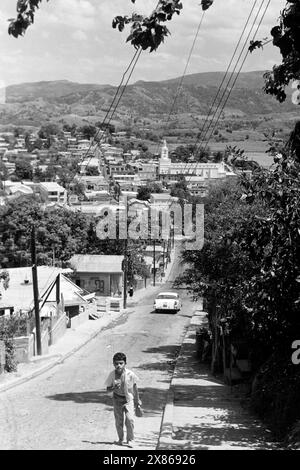 Ein Junge läuft die Straße Lorencita Ramirez in San Germán entlang, im Hintergrund ist die Pfarrkirche San Germán de Auxerre zu sehen, Puerto Rico 1966. A boy walks along Lorencita Ramirez Street in San Germán, with the parish church of San Germán de Auxerre in the background, Puerto Rico 1966. Stock Photo