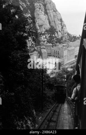 Blick auf den Marienplatz, den Hauptplatz der Klosteranlage von Montserrat aus dem Wagon der Zahnradbahn, die neben der Seilbahn Besucher und Gläubige zum Kloster und den Wanderrouten auf dem Berg bringt, Katalonien 1957. View of St Mary's Square, the main square of the monastery complex of Montserrat, from the carriage of the cog railway, which, in addition to the cable car, takes visitors and believers to the monastery and the hiking routes on the mountain, Catalonia 1957. Stock Photo