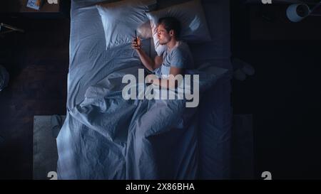 Top View Apartment Bedroom: Handsome Young Man Using Smartphone in Bed at Night. Guy Browsing Through Social Media, Using Dating Apps, Remote Work Software, Internet Online Shopping on Mobile Phone Stock Photo