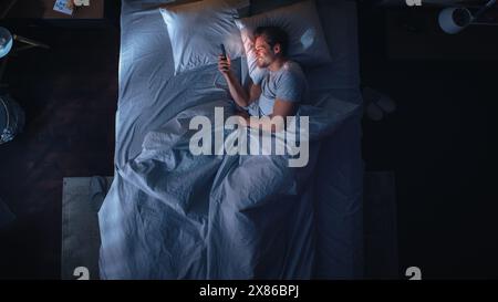 Top View Apartment Bedroom: Handsome Young Man Using Smartphone in Bed at Night. Smiling Guy Browsing Social Media, Dating Apps, Doing Internet Online Shopping on Mobile Phone. Top Down Stock Photo