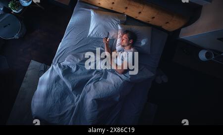 Top View Apartment Bedroom: Handsome Young Man Using Smartphone in Bed at Night. Smiling Guy Browsing Social Media, Dating Apps, Remote Work Software, Doing Internet Online Shopping on Mobile Phone Stock Photo
