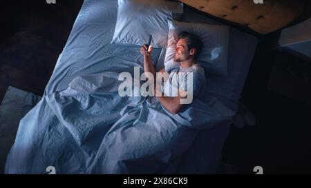 Top View Apartment Bedroom: Handsome Young Man Using Smartphone in Bed at Night. Smiling Guy Browsing Social Media, Dating Apps, Remote Work Software, Doing Internet Online Shopping on Mobile Phone Stock Photo