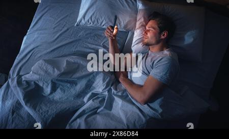 Top View Apartment Bedroom: Handsome Young Man Using Smartphone in Bed at Night. Smiling Guy Browsing Social Media, Dating Apps, Remote Work Software, Doing Internet Online Shopping on Mobile Phone Stock Photo