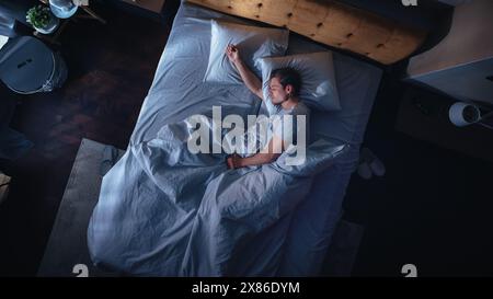 Top View Apartment Bedroom: Handsome Young Man Sleeping Cozily on a Bed in His Bedroom at Night. Comfortable Apartment with Guy Resting for Productive next Day. Top Down Above Stock Photo