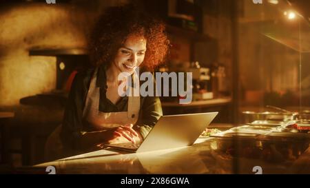 In Restaurant: Beautiful Female Chef Using Laptop Computer. Authentic Pizzeria With Delicious Organic Eco Food. Bi-racial Female Entrepreneur Working in Her Small Business Family Shop Stock Photo
