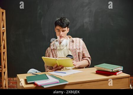 Young man, student sitting at desk and involved on book reading. Chalkboard background. Sitting in classroom and studying Stock Photo