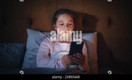 Small Girl Using Smartphone in Bed Before Going to Sleep. Cute Kid Checking Social Media, School Schedule, Reading Homework on Digital Ebook, Chatting with Friends Online. Stock Photo
