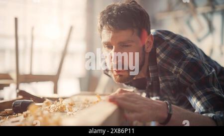 Handsome Young Artisan Craftsman in Checkered Shirt Using Hand Plane to Shape a Wood Bar. Carpenter Working on a Project in a Loft Studio with Tools on Walls. Stock Photo