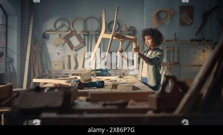 Young Female Carpenter Reading Blueprint, Making Notes, Starting to Assemble Parts of a Wooden Chair. Stylish African American Furniture Designer Working in a Studio in Loft with Tools on Walls. Stock Photo